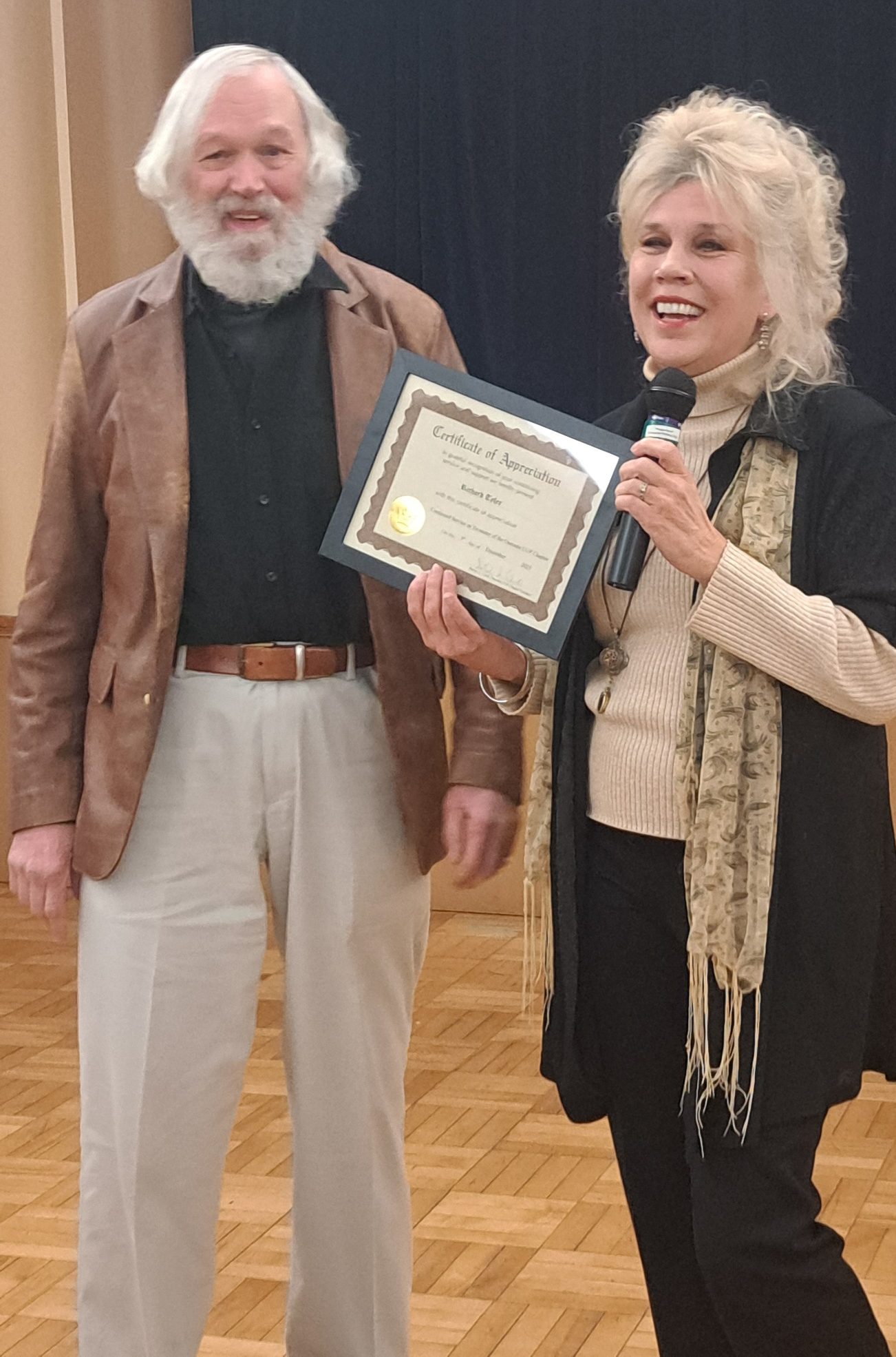 Richard stands next to Shirley at the UUP Oneonta 2025 Holiday Gathering. They are smiling mirthfully as Shirley presents Richard with a certificate.